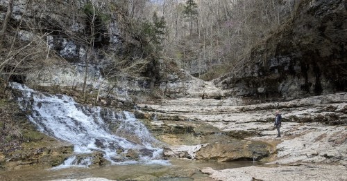 A serene landscape featuring a waterfall cascading over rocky terrain, surrounded by trees and steep cliffs.