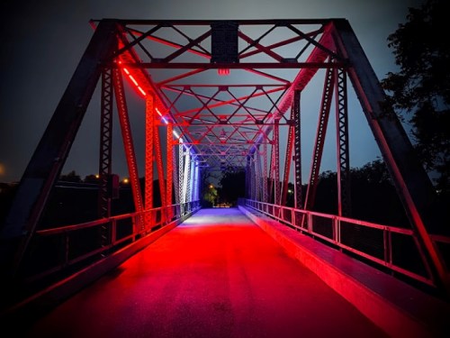 A lit metal bridge at night, featuring red and blue lights illuminating the structure and pathway.