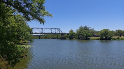 A calm river scene with a bridge in the background, surrounded by lush greenery under a clear blue sky.