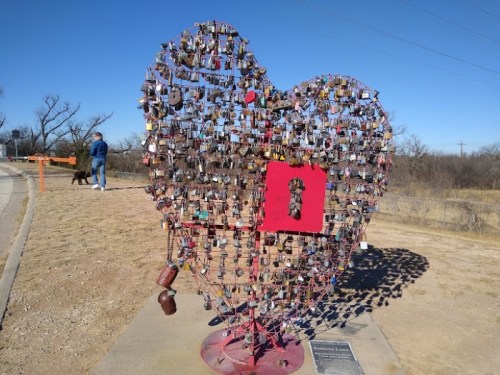 A heart-shaped sculpture made of locks, with a red center, set against a clear blue sky and a person walking nearby.