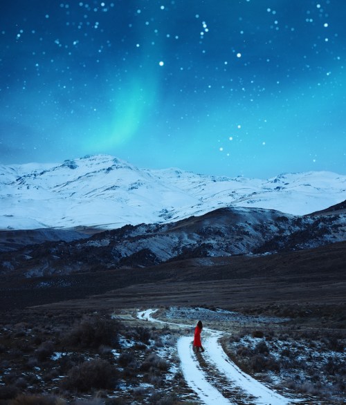 A woman in a red dress walks along a snowy path under a starry sky and northern lights over snow-capped mountains.