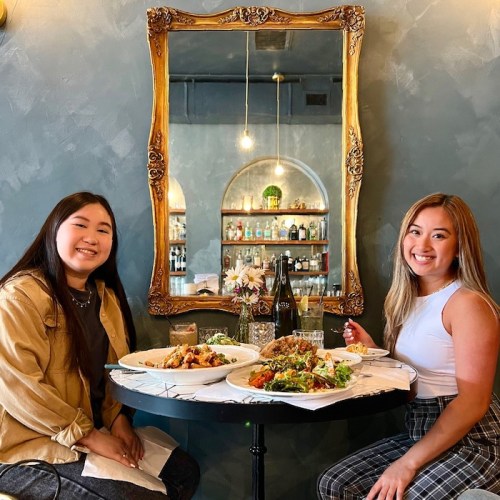 Two women sit at a table with a variety of dishes, smiling in a stylish restaurant with a decorative mirror.