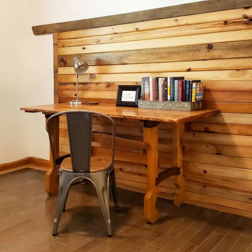 A wooden desk with a chair, lamp, and books against a rustic wooden wall.