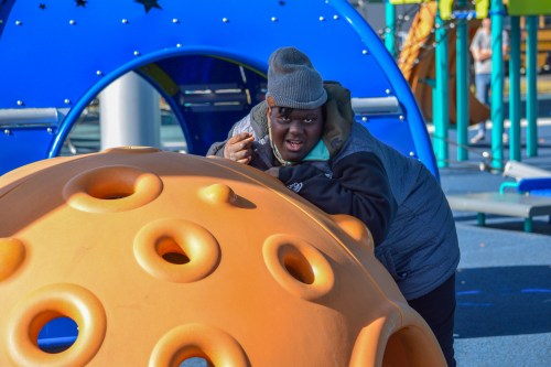 A child in a gray beanie plays on an orange playground structure, smiling and having fun in a colorful play area.