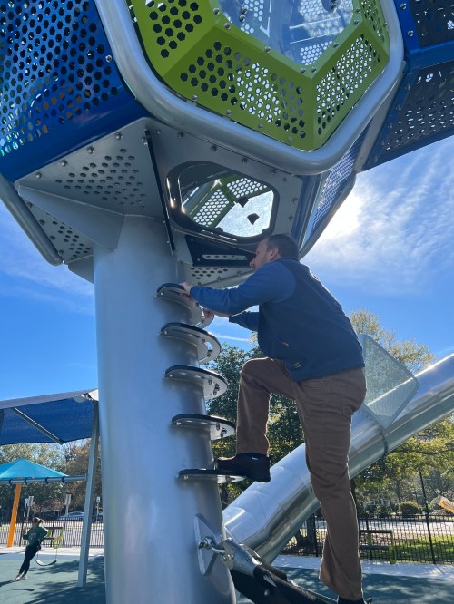 A person climbs a modern playground structure on a sunny day, reaching for the next step.
