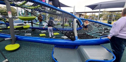 Children play on a colorful, modern playground structure with nets and climbing features under a shaded area.