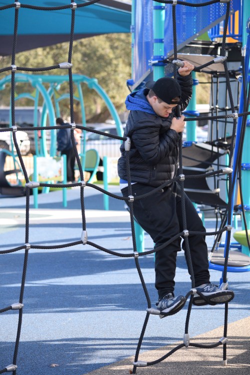 A person climbs a rope structure at a playground, wearing a black jacket and cap, with colorful play equipment in the background.