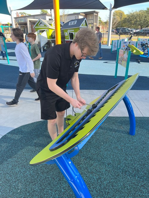 A young man in a black shirt interacts with a colorful play structure in a playground, with children in the background.