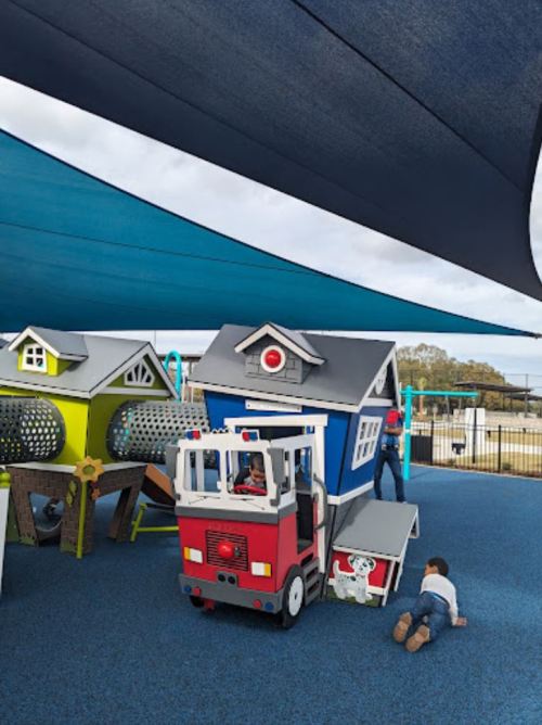 Colorful playground with a fire truck and playhouses, shaded by a large canopy, children playing nearby.