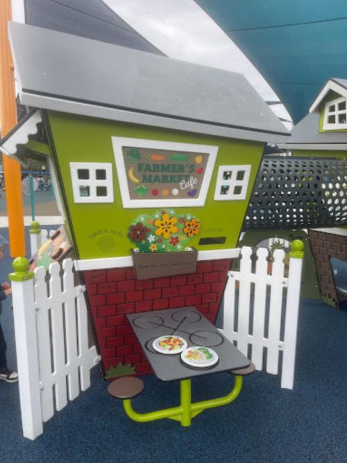 Colorful play structure resembling a farmer's market café with a table and plates, set in a children's play area.