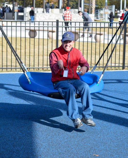 A person in a red vest sits on a blue swing, smiling, in a playground with people in the background.