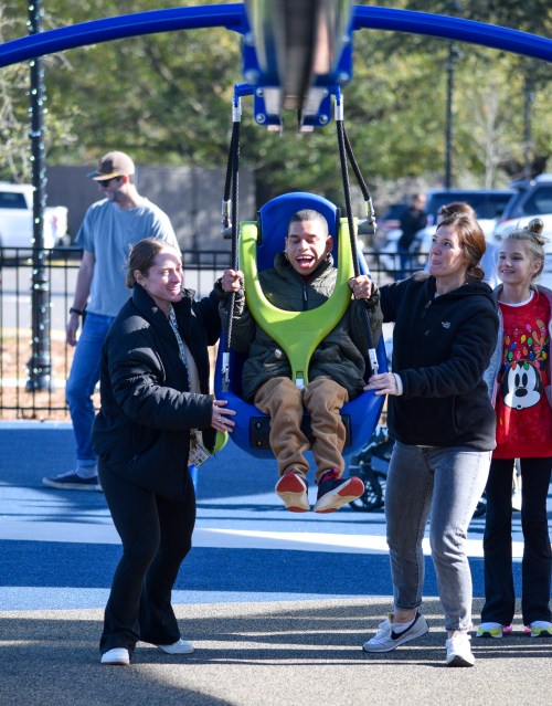 Two adults assist a child in a swing at a playground, with other children and adults in the background.