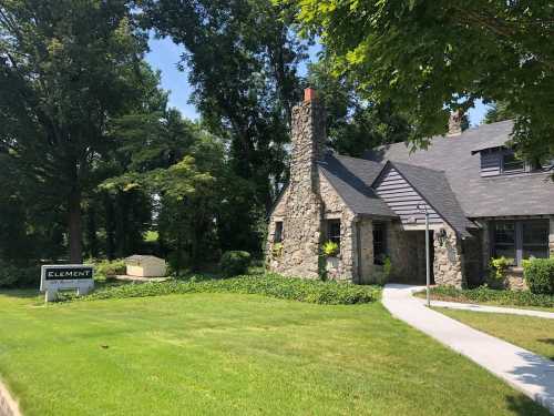 A stone house with a chimney, surrounded by greenery, next to a sign that reads "ELEMENT." Bright, sunny day.