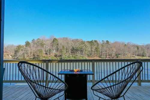 Two chairs facing a calm lake with a small fire pit on a wooden deck, surrounded by trees under a clear blue sky.