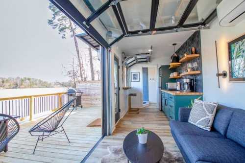 Cozy interior of a modern cabin with large windows, a sofa, and a kitchen, overlooking a scenic outdoor deck.