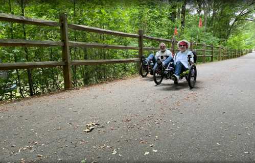 Two people riding recumbent tricycles on a tree-lined path, with a wooden fence in the background.