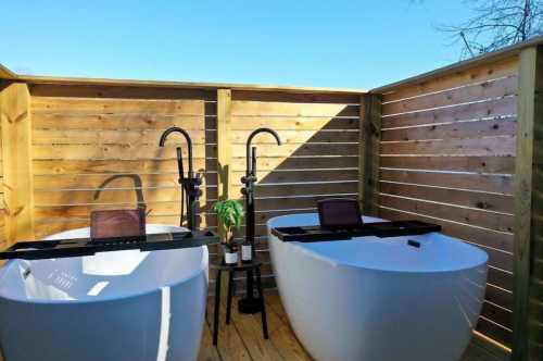 Two modern bathtubs on a wooden deck, surrounded by a wooden fence, with a small table and plants nearby.