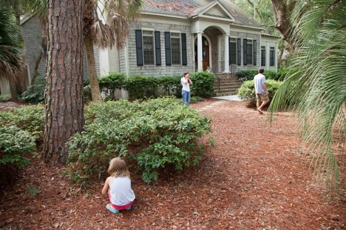A family explores a garden with a house in the background, surrounded by trees and shrubs. A child sits on the ground.