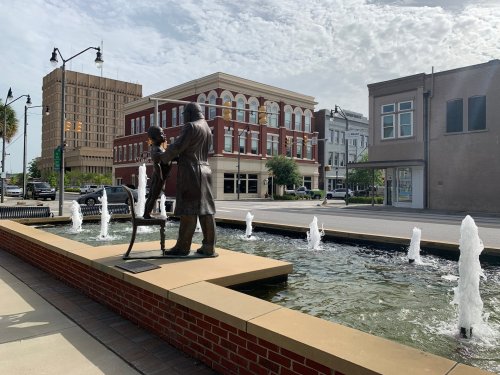 A statue of a man and woman stands by a fountain in a city street, with buildings and a cloudy sky in the background.