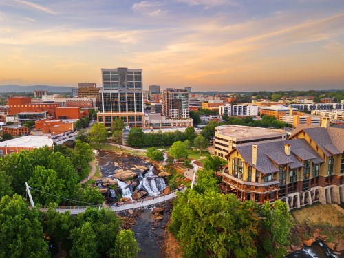 Aerial view of a cityscape featuring buildings, greenery, and a waterfall, with a sunset in the background.