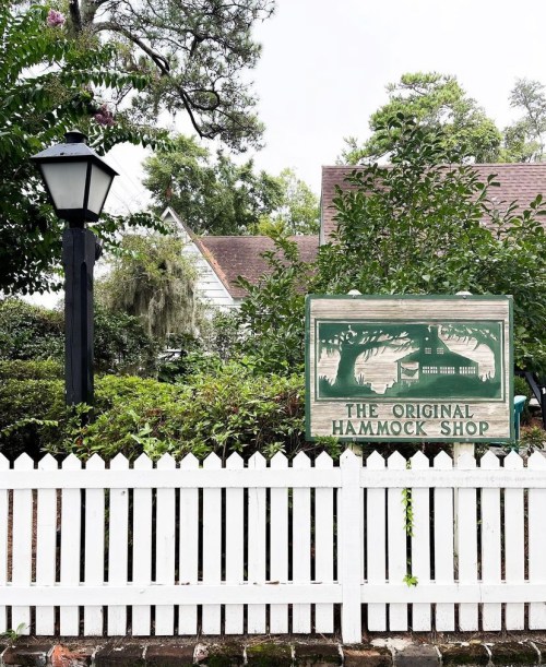 Sign for "The Original Hammock Shop" beside a white picket fence and a lamp post, surrounded by greenery.