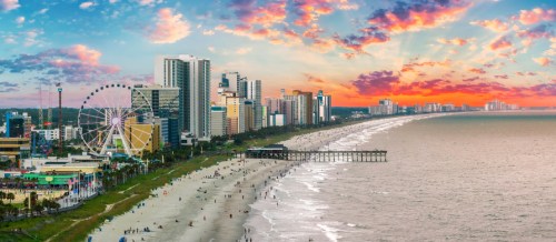 A vibrant sunset over a beachside city, featuring high-rise buildings, a pier, and a Ferris wheel along the shoreline.