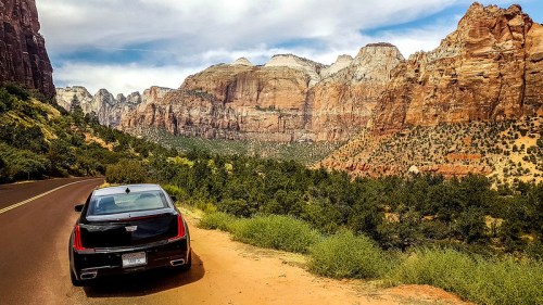 A black car parked on a scenic road with towering red rock formations and green vegetation in the background.