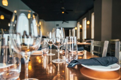 A dining table set with various glassware and black napkins, illuminated by soft pendant lights.