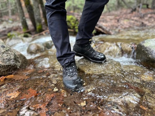 A person stands in a stream, wearing black hiking boots and pants, with water splashing around their feet.