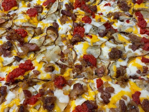 Close-up of a pizza topped with cheese, crispy potato slices, bacon, and dollops of red sauce.