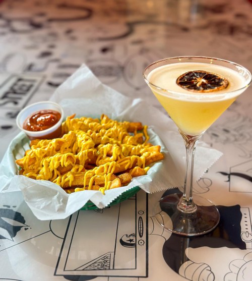 A plate of cheesy fries with a side of sauce, next to a yellow cocktail garnished with a dried lemon slice.