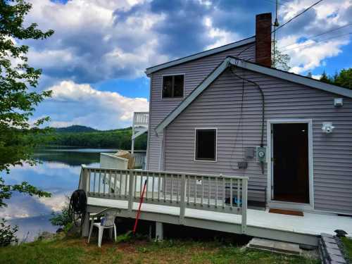 A lakeside house with a deck, surrounded by trees and reflecting clouds in the water.