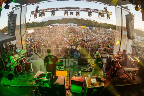 A lively outdoor concert scene with a large crowd enjoying a performance on stage, surrounded by trees and tents.