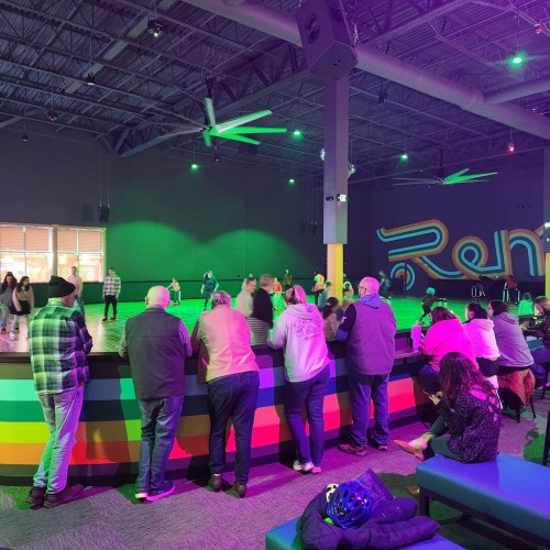 A vibrant indoor roller skating rink with people skating and a colorful bar area in the foreground.