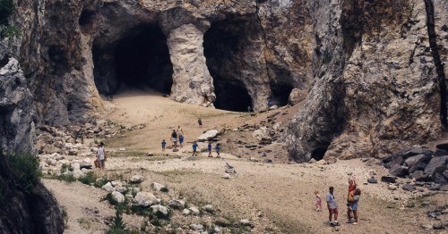 A group of people exploring a rocky cave landscape with two large cave openings and a sandy path.