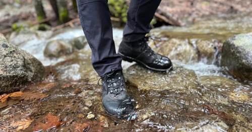 A person stands in a stream, wearing wet black hiking boots and dark pants, surrounded by rocks and flowing water.
