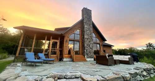 A cozy log cabin with a stone chimney, surrounded by nature, featuring a patio with seating and a colorful sunset sky.