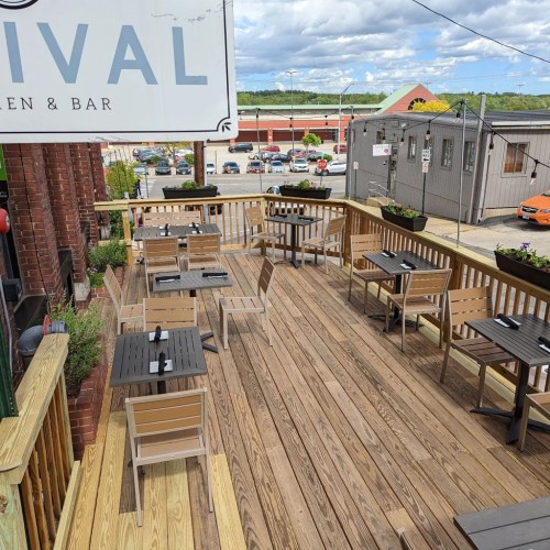 Outdoor dining area with wooden tables and chairs, overlooking a parking lot and distant hills under a cloudy sky.