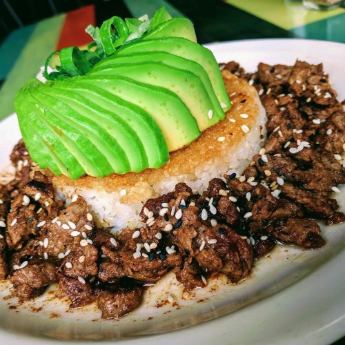 A plate of rice topped with sliced avocado and seasoned beef, garnished with green onions and sesame seeds.
