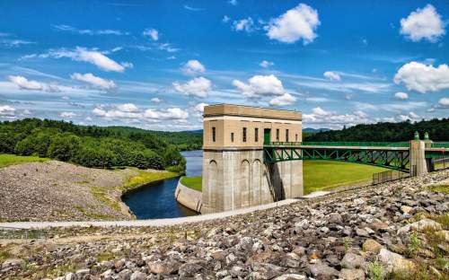 A scenic view of a dam with a control tower, surrounded by lush greenery and a blue sky with fluffy clouds.