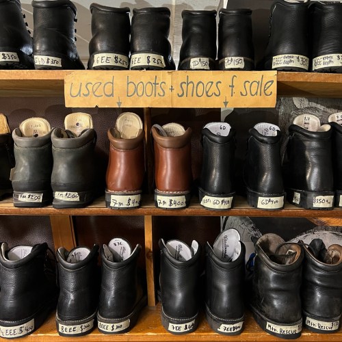 A shelf displaying various used boots and shoes for sale, labeled with prices and sizes.