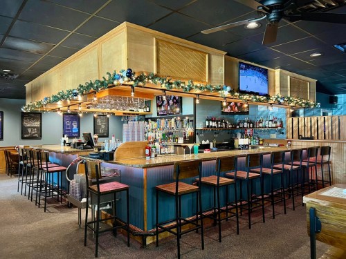A cozy bar with wooden accents, festive decorations, and a TV displaying a sports game. Bar stools line the counter.