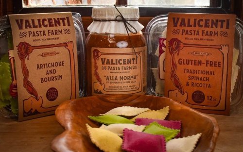 Three Valicenti Pasta Farm products: a jar of "Alla Norma" sauce, and two pasta packages, with a bowl of colorful ravioli.