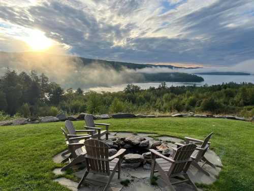 A serene lakeside view at sunrise, featuring a fire pit surrounded by wooden chairs and misty hills in the background.