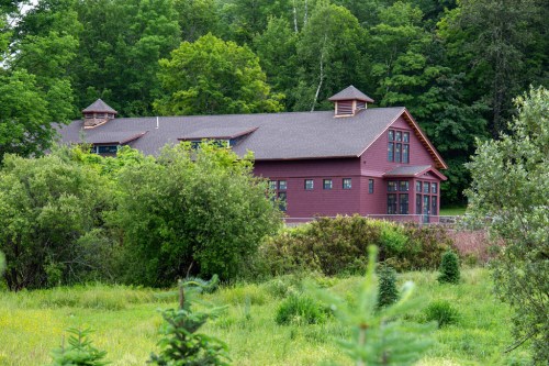 A large red building surrounded by greenery, set against a backdrop of trees and a grassy area.