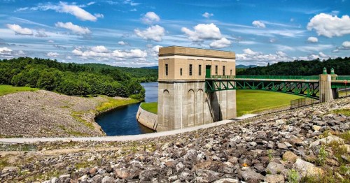 A scenic view of a dam with a control tower, surrounded by lush greenery and a clear blue sky with fluffy clouds.