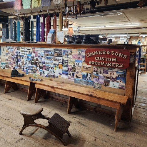 A wooden shop interior featuring a wall of photos, a sign for "Limmer & Sons Custom Bootmakers," and wooden benches.
