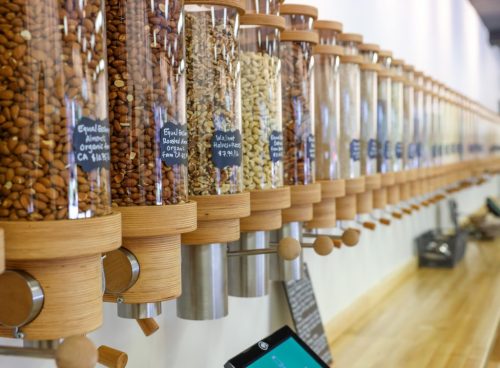 A row of clear dispensers filled with various nuts, labeled with prices, against a wooden countertop.