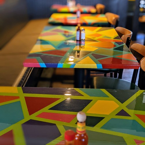 Colorful, geometric-patterned tables in a restaurant, with bottles of hot sauce on each table.