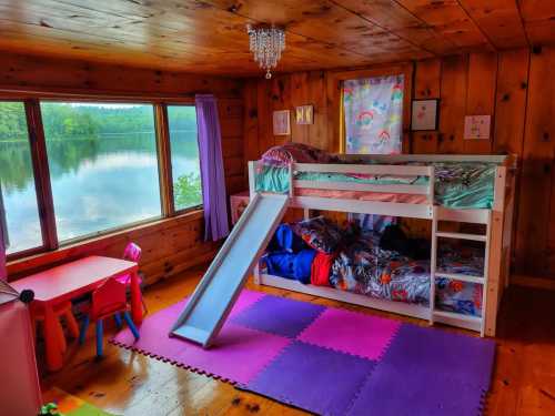 Cozy wooden room with a bunk bed, slide, colorful rug, and a view of a lake through large windows.